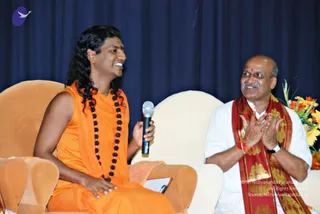 A photograph of two men seated on cream and orange upholstered chairs before a dark blue draped curtain at a stage event. On the left, a man with long, swept-back dark curly hair, a small red bindi, and an orange sleeveless robe with a long black beaded mala sits holding a microphone and smiling. Probable Swamiji by appearance cues: long hair, saffron clothing and rudraksha-style mala. On the right, an older man with a shaved head, grey moustache, white shirt and a red and gold shawl with a red wrist thread is clapping his hands. A bouquet of orange and yellow flowers stands at the right edge.