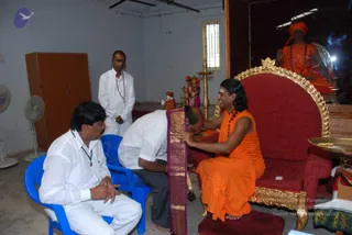 Indoor room with white walls and a window grille. A man with long dark hair, wearing an orange robe, rudraksha mala and bare feet, sits on an ornate red and gold throne-style chair smiling while a man in white shirt and dark trousers bends forward to touch his feet. A seated man in white shirt with a pendant sits on a blue plastic chair at left, and a standing man in white dhoti-style clothing with a mala stands behind. A purple and gold shawl is draped over the chair. A brass oil lamp, a small deity murti and a framed portrait in orange robes with a turban hang on the right wall, and an orange-robed figure with a mala is shown in the framed photo above the chair. A brass tray with offerings sits at the right edge. A wall fan stands at left.