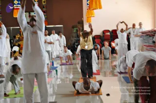 A yoga session in a hall with cream walls and a brown wall, decorated with orange and colourful hanging fabric panels. Many participants in white clothing stand, bend and stretch on mats across a polished reflective floor. In the foreground a man in white stretches forward on an orange mat, and a man in a gold shirt with dark trousers and a neck mala stands in a forward bend on an orange mat. Some people raise their arms overhead, others with hands joined above their heads. Red chairs line the back wall and a stack of folded blankets is visible at right. A yoga group practice is in progress.