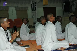 A seated group of young men in white kurta-style clothing sit on orange floor mats in a hall with cream curtains and a wall sign. The man at left has white vibhuti on his forehead, a rudraksha mala, a gold bracelet, and palms pressed together in prayer. Several others have shaved heads, glasses, and white clothing, and some sit with hands folded or palms joined. A small dark book lies on the mat in front. No teacher is clearly visible in the frame. Orange and white tones dominate, with a yellow hanging decoration at top.