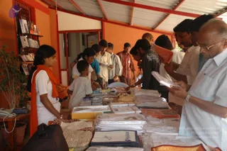 A bookstall or merchandise counter in an orange-walled building with a red-and-white corrugated roof. A young woman in white with a saffron-orange shawl stands at left behind a table covered with clear-wrapped bundles, folded cloth, CDs and booklets. Several men and women in casual and white clothing crowd the table, one older man at right in a pale blue shirt and glasses browses a booklet. Shelves of books and pamphlets line the back wall, and a potted plant and telephone sit at left.