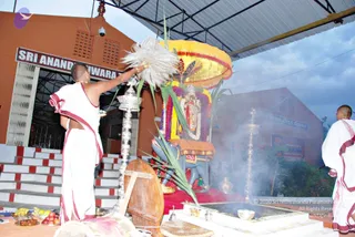 Outdoor temple scene under a corrugated metal canopy in front of an orange-walled building with a signboard reading Sri Ananda Eshwara. A priest in white dhoti with red borders and a bare upper body waves a large white feather fan over a silver lamp stand. Behind him stands a decorated deity murti on a vahana with a yellow canopy, garlanded with flowers and dressed in red and purple cloth. A second priest in white with red-bordered garments stands at the right. A fire pit with smoke rises in the foreground, and fruit offerings and brass vessels sit on the left.