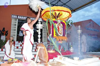 Outdoor temple scene at dusk with a priest in white dhoti and red-bordered cloth raising a white feather fan (chamara) over a richly decorated deity murti. The murti is seated on a blue mount with a yellow and red canopy, garlands of purple and yellow flowers, and banana plant leaves on either side. Smoke rises from a fire pit in front, with silver oil lamps and brass vessels on a platform. Another white-clad priest stands at the right and a bare-chested priest with a gold necklace sits at the left on red-and-white steps. Orange-walled buildings with signboards stand behind.