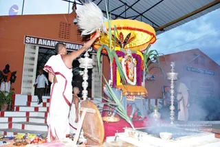 Outdoor temple ritual at dusk under a corrugated metal canopy. A priest in white dhoti with red-bordered cloth, bare-chested, raises a white chamara fan over a decorated deity murti seated on a blue vahana-style figure beneath a large yellow and red umbrella-like canopy and a garland of purple and white flowers. Silver oil lamps stand on either side and smoke rises from the fire pit in front. Offerings of fruit and brass vessels sit on the ground in the foreground. Another white-clad priest stands near a tiled wall at right and a figure in a saffron-trimmed cloth appears in the background. A signboard reads Sri Anandeshwara in Kannada script on the building. Shaved-headed priests perform puja.
