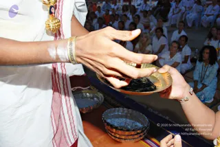 Close photo of a ritual exchange in an ashram hall. In the foreground a figure in white clothing with a red-and-white striped border and a gold pendant with a small portrait medallion holds out a brass plate containing dark, thick substance. Another person's hand, with a silver bracelet, receives the plate. Behind, several stacked dark metal bowls rest on an orange and blue cloth. The seated audience wears white and light blue clothing and sits in rows on a hall floor.
