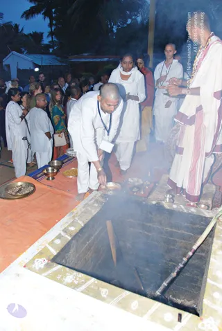 Outdoor fire ritual at dusk with a square homa pit lined with stones and smoking, edged with white chalk-drawn patterns. A man in white clothes with a blue lanyard bends over the pit, holding a small brass dish of offering. Behind him a woman in white with a white shawl stands with hands folded. On the right, a figure in white robes with maroon and cream borders stands at the pit's edge with a long rudraksha-style necklace, while a bald man in white with a mala looks on. Brass bowls, a plate of grain, and a tray of offerings sit on an orange cloth. A crowd of people in white and colourful clothes stands behind under palm trees and tin-roofed huts.