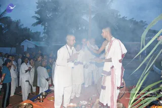 Outdoor photograph at dusk of a ritual gathering with thick smoke rising from a fire pit in the foreground. A bald priest in white dhoti and a white shawl with red border, wearing a rudraksha-style bead necklace and a tilak on his forehead, stands at right holding his hands toward a man in white kurta who stands with palms pressed together at left. Behind them, several men in white clothing stand in the smoke, and a crowd of devotees in white and other colours stands beneath palm trees. Brass vessels, bowls and oil lamps sit on a mat in front, and a green palm frond is visible at the right edge. The priest's figure shows no distinctive a person cues beyond ordinary white ritual clothing, so identity is unsure.