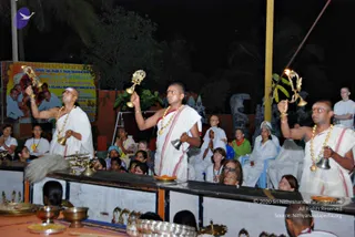 Night outdoor ritual at a temple-style courtyard. Three priests in white dhotis with pink-red borders and white shawls stand at a raised platform, each wearing ornate gold-beaded necklaces. The two on the left and right raise lit oil lamps (aarti) high in their right hands, while the centre priest holds a handbell and a small bell in his hands. Brass pots, plates and lamps sit on the platform in front of them. Behind the platform a seated audience, including women in white saris and a woman in a green top, watches with hands folded in devotion. A banner with Kannada text and a photo of a saffron-robed figure hangs to the left, and stone sculptures and palm trees fill the dark background. This aarti ritual is consistent with the event; the page describes a Ganapathi homa album.