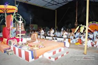 Night-time photograph of an open-sided pandal at an ashram, with a raised platform draped in orange cloth and striped red-and-white steps. On the platform, a priest in white dhoti and shirt stands before a homa-style setup of brass plates, flowers, fruit and a conch-like object. Another white-clad priest holds a ritual item nearby. Rows of seated devotees in white, a few in saffron, sit on mats facing the platform. A framed deity image hangs at left beside a tall brass lamp stand and a red-and-gold canopy. Several dozen people are visible in the scene. The picture is taken from the side of the pandal.