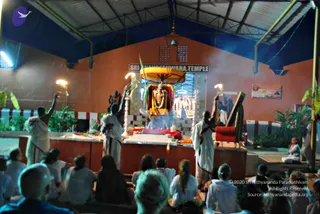 Night ceremony in an open-sided hall before a shrine labelled "SRI RUDRAKSHESHWARA TEMPLE" in dark lettering on a white board. A decorated deity murti sits under a yellow-and-orange canopy with marigold garlands and a flower-covered central form, with smoke rising. Three men in white dhotis stand on the platform raising lit lamps, one holding a long-handled lamp toward the deity. A seated figure in white sits at the right beside a red-cushioned chair. In the foreground, a crowd of devotees in white and light clothing sits facing the altar, some with shaved heads, others with long hair. Fluorescent lights and potted banana plants frame the scene, with a small framed notice on the rear wall.