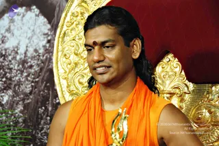 A man with long dark hair swept back sits before an ornate gold throne-like backdrop with a red panel behind it. He wears a saffron-orange sleeveless garment draped over the shoulders, a red tilak mark on his forehead, and a gold ceremonial pendant on his chest. A pale, misty mountain-like image and green leaves appear at the left edge. Swamiji (probable): long swept-back hair, saffron clothing and ornate gold ceremonial adornments suggest the presiding teacher, shown seated and speaking.
