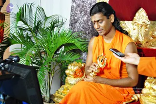 A photograph of a man seated on an ornate golden-and-red throne, wearing an orange saffron robe draped over his shoulders and a sleeveless orange garment, with a red tilak mark on his forehead and long dark hair swept back. He holds a small gold trident (trishul) in his hands and looks down at it. Beside him sits a gold lion-shaped figure, and a green potted palm stands at left. A person in orange clothing extends a hand holding a black mobile phone toward him from the right edge, and a camera and monitor appear at the left.