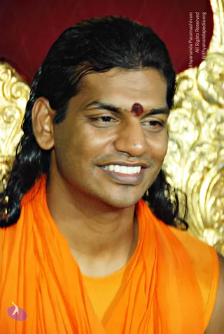 A close-up photograph of a smiling man with long, swept-back dark hair and a red bindi on his forehead, wearing a saffron-orange shawl wrapped over his shoulders. Behind him is ornate gold carved decoration against a deep red backdrop. Based on the appearance cues of long swept-back hair, saffron clothing and the ornate gold setting, this is. The page's album title and excerpt reference a morning message on revival of Vedic tradition.