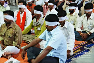 A photograph of a seated group of men in a meditation or workshop hall, all wearing white cloth blindfolds tied over their eyes. Several sit cross-legged on orange floor mats and dark patterned rugs, dressed in pale blue, white, yellow-green, beige and striped shirts and dhotis. One man in front wears a light blue shirt and dark trousers with his hands resting on his knee. Another on the left wears a bright orange scarf over a white shirt. Some have name badges clipped to their shirts. Chairs are visible at the back of the room.