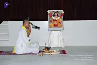 A photograph of a shaved-headed man in white ritual clothes with a yellow shawl sitting cross-legged on a red patterned rug, hands folded in anjali mudra before a microphone on a stand. In front of him are brass puja items including a small oil lamp with a lit flame, a bell, a brass pot and a bowl on a tray. Behind them on a draped white table stands a framed portrait labelled "PARAMAHAMSA NITHYANANDA", showing a figure in saffron robes seated in meditation, garlanded with white and red flowers. A dark red curtain and white wall form the backdrop.