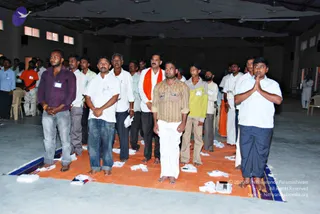 A photograph of a large indoor hall with a grey concrete floor and ceiling fans. Men stand in rows on an orange tarpaulin, many barefoot, with white cloths and small items laid on the floor. In the foreground, a man in a purple shirt and jeans stands with hands clasped, a man in a white shirt and white trousers beside him, and a man in a white shirt and orange shawl in the centre. The man at centre right wears a striped brown shirt and white dhoti, and a young man on the right wears a white shirt and dark trousers with palms pressed together in namaskar. Other men in white shirts and some in orange cloth stand behind them. Plastic chairs sit along the left wall, and a small white-clothed figure stands at the far right edge.