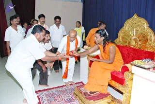 Indoor photo of a seated figure on an ornate gold-and-red throne receiving an offering. A devotee in a white shirt and dhoti kneels on a red and blue patterned rug, extending both hands to offer a red fruit or sweet. Another man in white with an orange shawl stands nearby with hands joined, and several men in white shirts and dhotis stand behind in a blue-curtained hall. A tray of sweets and flowers sits on a side table at right. The setting resembles a satsang or darshan hall.