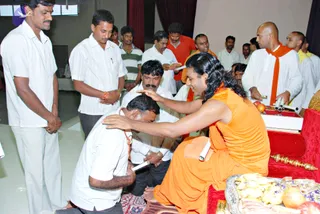 A seated figure in orange robes with long, swept-back dark hair and a rudraksha mala places his hand on the head of a kneeling man in a white shirt who bows his head. The robed figure sits on a red cushioned throne-like seat with a gold ceremonial staff beside him. Around them, men in white shirts and white or orange garments stand and watch in a hall. A silver tray holding fruit and offerings sits in the foreground, with a red box and rolled cloth papers nearby. The appearance matches Swamiji (probable), with long hair, orange clothing and rudraksha beads, and he is blessing the devotee.