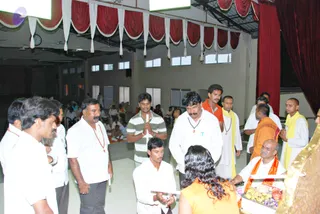 Indoor hall with red and white draped valances and a red curtain along the right side. Several men in white shirts and saffron or yellow shawls stand and kneel near the centre; one man in the foreground with folded hands holds a small white item. A man in an orange-saffron robe with a yellow-orange shawl sits at the right, beside a tray of flowers and offerings. A woman in an orange top faces the tray from the foreground. Rudraksha malas are visible on several men. Seated people fill the background. a person is not clearly identifiable from appearance, so no candidate is asserted.