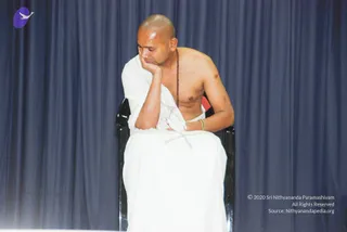 A photograph of a shaved-headed man seated on a black chair against a dark blue draped curtain. He is bare-chested, wearing a white dhoti and a white cloth draped over his left shoulder, with a red-brown rudraksha mala hanging on his chest and a gold bangle on his wrist. His head rests on his right hand, his elbow on his knee, as he looks downward in a thoughtful pose.