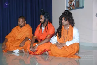 Three men sit cross-legged on a glossy tiled floor in orange saffron robes, reflected in the surface. The man on the right wears a white inner kurta, a rudraksha mala, a shawl over his shoulders, and long curly dark hair piled in a topknot; his hands rest together in his lap. The man in the center has long straight black hair, a saffron wrap with red-gold border and a gold bangle and wristwatch, looking to the side. The man on the left has short hair, closed eyes and a plain orange wrap, sitting barefoot. A blue curtain hangs behind them and a person with a microphone is visible at a window.