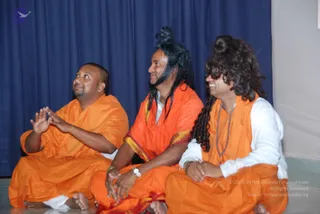 A photograph of three men seated cross-legged on a floor in front of a dark blue curtain. The man on the left, with short hair and beard, wears plain orange robes and raises his palms, gazing upward. The middle man has long black hair tied in a topknot, wears an orange robe with a gold-bordered edge and a white undergarment, and wears a wristwatch. The man on the right has long wavy dark hair, wears an orange shawl over a white top, and a rudraksha mala around his neck. A grey wall with a small white sign is visible at right.