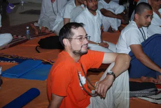 A photograph of a seated group on an orange floor covering during a satsang-style gathering. In the foreground a bearded man in glasses wears an orange t-shirt and white pants and holds a dark rudraksha-style mala beaded necklace, looking to his side. Behind him men in white clothing sit cross-legged or on the floor; one has a short-cropped head and a dark cord necklace, another wears a white collared shirt. A blue rolled mat, a water bottle and dark bags lie on the floor, with a striped mat at left. No deity, throne or a person depiction is visible.