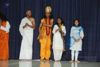 A photograph of a stage performance in front of a dark blue curtain. A performer in a golden-yellow dhoti and matching shawl, wearing a gold crown and a red and white flower garland, stands centre with arms folded across the chest. To his left, a bare-chested man in a white dhoti and shawl with a sacred thread smiles. To the far left, a partly visible figure wears an orange robe. To the right, a woman in white with a patterned shawl holds a yellow object, and a woman in white with a blue dupatta stands beside her. Four to five people are visible, all barefoot on a glossy stage.