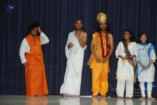 Five people stand in a row on a stage before a dark blue draped curtain, appearing to pose for a group photo during a dramatised skit. On the far left, a man in a white shirt and long saffron-orange dhoti or wrap, with swept-back dark hair, touches his head with one hand. Next, a bald man bare-chested in a white dhoti and shawl gestures with one hand. Third from left, a man wears a golden crown, a red and yellow flower garland, a golden-yellow garment and bangles, holding his hands together at his waist. Two women stand on the right: one in white with a patterned brown shawl holding green and yellow props, and one in white with a light blue sheer scarf.