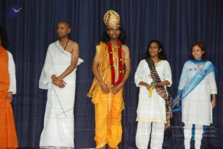 A photograph of a stage with a dark blue draped curtain backdrop showing several people standing in a row. In the centre, a man wears a tall gold crown, a red and yellow flower garland, and an orange-gold dhoti and shawl, with gold armbands. To his left, a bald man in white draped cloth stands with his hands clasped. To the right, a woman in a white kurta and white trousers holds two yellow sticks and wears a dark patterned shawl, and beside her a smiling woman in white with a light blue dupatta. At the far left edge, a partly visible person in orange robes stands.
