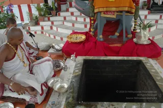 A photograph of a temple courtyard with a square stone-rimmed yagna pit filled with dark ash or embers at the lower right. Behind it, a grey bull (Nandi) murti sits on a red-draped platform with an orange-and-gold cloth over its back, garlanded with marigold and white flower strings, and a silver vessel with a leafy plant stands beside it. On the left, a bald priest in white dhoti and shawl with red borders and a gold necklace sits cross-legged on the floor, with a second person in white partly visible behind him. Silver plates, bowls and a small silver kalasha sit on the floor near the fire pit, and a microphone stand is positioned at left. Red and white striped steps lead up in the background.