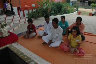 A photograph of five people seated cross-legged on an orange cloth outdoors in front of a temple-like area with red-and-white striped steps and a dark stone platform. In the foreground a girl in a yellow top and pink skirt sits with hands folded, a vermilion mark on her forehead and a name badge on her chest. Beside her a young boy in a white kurta sits with eyes closed, next to another boy in white and a woman in a teal top and glasses. At the far right a man in a salmon-pink shirt sits with hands on his knees. At the left edge a girl in a maroon top sits on a blue mat beside a silver tray with flowers and a brass vessel, and a dark stone deity figure with a garland is partly visible at the top left. Red banners and green shrubs fill the background.