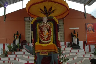 A decorated deity murti of a Hindu god stands on a grey elephant-shaped vehicle in front of an orange-walled building with a sign reading Sri An. The figure wears a crown, gold ornaments and a red-and-gold garment, and is draped in layered garlands of red, orange, yellow and white flowers. A large yellow umbrella with a lotus-motif canopy shades it, and black stone guardian statues flank the steps, each garlanded with orange and white. To the right a poster shows a smiling man in an orange turban and saffron robes labelled Paramahamsa Nithyananda, with text reading Every Saturday and Sunday. The steps are painted red and white.