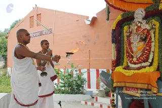 Outdoor temple photograph of an aarti being offered to a Ganesha murti decorated with garlands of orange, yellow and red flowers and silver ornaments. Two shaved-headed priests in white dhotis with red borders and gold necklaces stand before it; the front priest holds a silver lamp with a flame in his right hand and a bell in his left, while the second, behind him, chants with a hand raised. A peach-coloured brick wall with a sign reading 'गणेश्वर मंदिर' (Ganeshwar Mandir) stands behind them, with green foliage and marigolds at left. A saffron cloth covers the base of the murti.