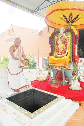 A shaved-headed priest in white dhoti and shawl with red borders stands at left, pouring offerings from a metal bowl toward a square black pit lined with white border. Beside him, a ganesha deity murti sits on a grey stand draped in orange cloth, decorated with garlands of marigold and red flowers and an ornate silver crown, under a large yellow umbrella canopy with dark decorative motifs. A red cloth covers the floor, with a brass plate, a silver pot wrapped in flowers and a brass tray nearby. A signboard in Devanagari reads Shri Anandeshwar temple. The priest wears a gold necklace and a sacred thread, and the scene suggests a homa or yagna ritual.