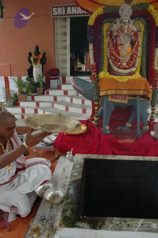A photograph of a temple shrine showing a silver-clad Ganesha murti seated on a grey mouse vehicle, draped in garlands of red, yellow and purple flowers beneath an orange canopy. In the foreground, a bare-headed priest in white dhoti and shawl with red borders, wearing gold necklaces and bangles, sits cross-legged holding a brass plate. Silver bowls and a small oil lamp sit on the orange floor near a black pit with a stone rim. Behind, a dark statue in white dhoti and orange shawl stands by red-and-white striped steps, and a red chair and potted plants are visible beside a doorway with a sign reading SRI ANAN.