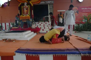 Outdoor photo at a temple-style courtyard. On a raised platform draped in orange cloth with a purple-and-white border, two people in yellow and red clothing lie prone in a deep forward bend, foreheads near the cloth, bare feet visible. A standing man in a white kurta and white dhoti watches beside a child in a red top near a stepped white-and-red staircase. Behind them stands a decorated deity murti of a seated figure on a blue mount, garlanded with flowers and an orange canopy above, with a shorter dark statue on the right. Brass bowls sit on the platform's left edge. A red banner on the right shows a smiling man in orange headwear and text reading Life Bliss Program, 2 Days Meditation Program, Physical Health, Mental Health, Radiance and a Sunday schedule.
