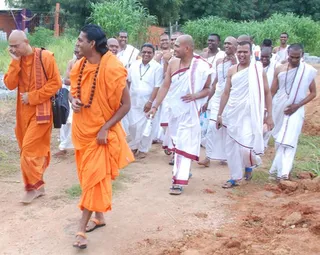 A group of men walks along a dirt path past green shrubs and a brick building. In the foreground, a man in saffron-orange robes carries a black bag and wears a rudraksha mala, looking down while smiling. Beside him, a man in saffron robes with long swept-back hair and a beaded mala walks with a light stride. Behind them, a row of men in white dhotis and shawls with red borders, many with shaved heads and beaded necklaces, walk and laugh together, some wearing sandals. Several men in white shirts and trousers walk among them.