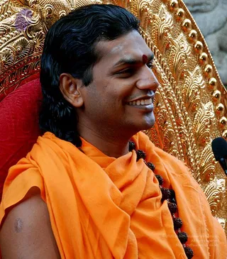 A smiling man with long dark swept-back hair and a red tilak on his forehead sits on an ornate gilded throne with a red cushioned backrest and carved floral filigree. He wears an orange saffron shawl over his shoulder and a dark rudraksha mala draped across his chest. A microphone stands at the right edge of the frame. Swamiji (probable): the long swept-back hair, saffron clothing, rudraksha mala and gilded throne match cues for the page's named teacher, though the identification is appearance-based.