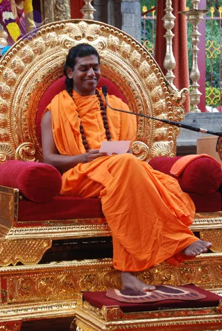 A man sits on an ornate gold throne with red cushions, smiling while reading a small pink paper. He wears an orange saffron wrap over his shoulder and body, a dark rudraksha mala around his neck, and a red tilak on his forehead. A microphone on a stand is near him, and gold lamp stands and garlands decorate the background. He is barefoot, resting his feet on a red cushioned footstool.