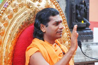 A man seated on an ornate gilded throne with a red cushioned back, raising his right hand in a gesture of blessing or teaching. He wears a saffron-orange garment draped over his shoulders, a rudraksha mala around his neck and a red tilak on his forehead, with long dark hair swept back. A microphone stand is visible at right beside a dark stone carved shrine or temple wall with a sculpted figure. Swamiji (probable) appears as the seated speaker, identified by long swept-back hair, saffron clothing and rudraksha mala. Marigold-like yellow flower visible near the carving.
