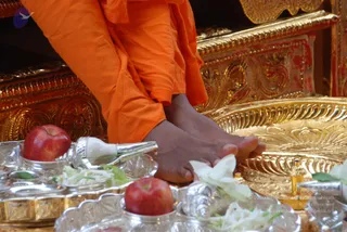 Close-up photograph of bare feet resting on a golden ornate platform during a padapuja ritual. The person wears orange saffron robes covering the legs. Around the feet are silver offering bowls holding red apples, white flowers and green leaves, a silver vessel with a white handle, and a small brass oil lamp. The golden carved throne and gilded tray frame the scene.