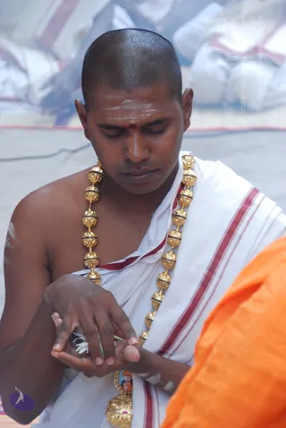 A shaven-headed man in a white dhoti with red-striped border sits with eyes lowered, holding white flowers and a small object cupped in his hands. He wears a heavy gold bead necklace and white sacred ash on his forehead with a red dot. Beside him, a figure in orange robes is partly visible at the right edge, and a small pendant on the gold necklace shows a figure in orange. The setting is a ritual gathering with white cloth in the background.