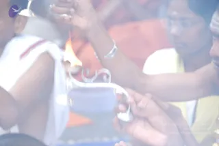 A blurred, washed-out close-up of a ritual scene with a lit flame at left and a silver ritual vessel with a curved handle at center. A hand holding a silver item rises from the upper right, next to a person in white and a figure in orange and white draped clothing. Another hand grips a beaded strand near the bottom. Visible people include a child in white at left, a woman with glasses and a bindi-marked forehead at upper right, and a partially visible person in yellow. The scene suggests a puja or aarti with a fire lamp. No one can be confidently identified as a person from appearance.