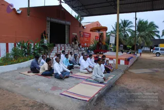 A photograph of devotees seated in rows on mats outside the Sri Anandeshwara Temple, a terracotta-orange building with a dark doorway and a sign reading SRI ANANDESHWARA TEMPLE. About twenty men sit on red-and-white striped steps and on woven mats on a tiled platform, many in white shirts and dhotis or trousers, some in light blue or beige shirts, one wearing a cap. A deity murti stands under a small orange canopy near the steps, garlanded with flowers. A banner reading Life Bliss Program hangs on the wall, with a portrait of a saffron-robed figure. Palm trees, marigold plants and a corrugated roof frame the scene under an overcast sky.