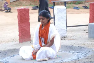 A woman sits cross-legged on a mat on bare ground, eyes lowered, hands resting in her lap. She wears a white dhoti-style garment with cream border, an orange-saffron shawl draped over her shoulders and across her chest, and a rudraksha-style mala. A small red bindi marks her forehead and her dark hair is pulled back. Around her, the ground shows grey ash, red and white stone pillars stand nearby, and sandals lie scattered in the background beside a figure in dark trousers walking past.