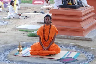 A man in saffron-orange robes sits cross-legged in meditation on a striped woven mat set on a circular concrete platform surrounded by grey ash. He wears a long rudraksha mala, has a red tilak on his forehead and eyes closed. Beside him stands a brass kalasha-style vessel. Behind him is a large orange stepped stone pedestal topped by a dark deity murti decorated with flower garlands. In the background, several people in white and yellow clothing sit on mats on the ground in meditation, with red signs in front reading about a sampling process.