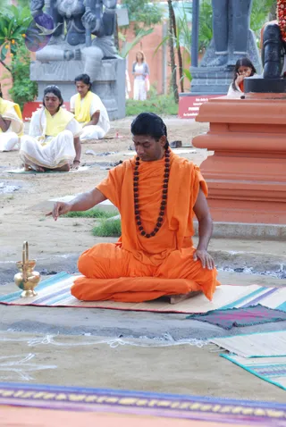 A man in orange saffron robes and a long rudraksha mala sits cross-legged on a striped mat outdoors, with his right hand extended toward a small brass vessel with a handle at left. His hair is swept back and he has a red mark on his forehead. Behind him, several women in white and yellow shawls sit on mats near grey stone deity sculptures and an orange pedestal. A woman in a grey top stands in the background among palms and plants, and a woman in orange touches her face at right. Charred ground and white kolam-like lines are visible.