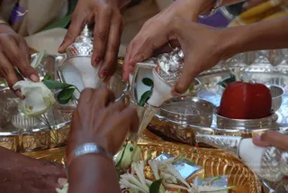 Close-up of hands pouring liquid from two silver-lidded white conch-shaped vessels over a gold tray during a ritual. A woman's hand with red-painted nails and a silver bangle holds a white rose. Silver bowls, a red apple, white jasmine-like flowers and green leaves lie on the tray. Other hands in the frame hold silver implements. The setting suggests an abhishekam offering with flowers and fruit.