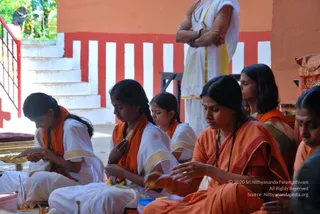 A photograph of a ritual scene on a covered temple-style platform with red and white striped walls. Several women sit cross-legged on the floor in the foreground, wearing saffron-orange shawls and white or cream garments with gold borders. The woman in the front right wears an orange sari with a rudraksha-style bead necklace and a red bindi, and she is handling items with her hands. Another woman in the centre wears a white outfit with an orange shawl and bends over a brass plate. Behind them a standing figure in white dhoti and shawl with a gold border stands near a staircase and red railing, arms folded and wearing beaded necklaces. Small brass vessels and blue and white containers sit on the floor, and a cloth-wrapped object rests on the right.